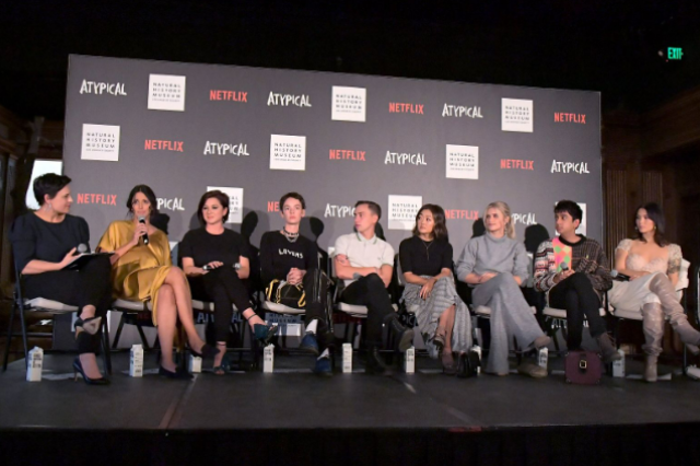 Row of people on a stage with one holding a microphone with the Atypical television show and Netflix logos repeated on a mural panel behind them.
