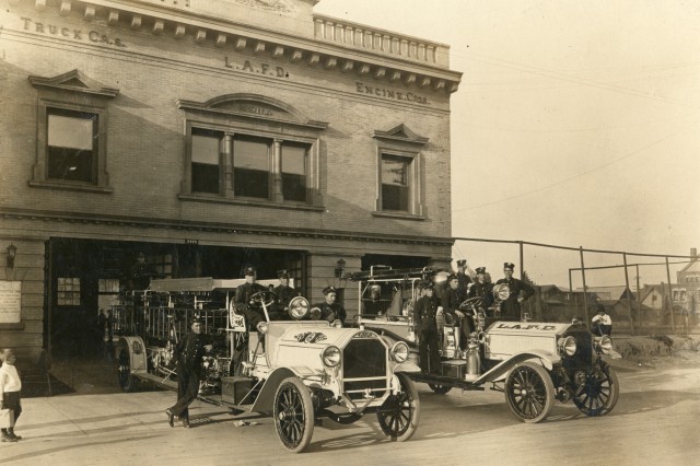 Antique photograph of two fire trucks in front of a brick two-story firehouse labeled LAFD. Five fire firefighters sit on the truck on the right. Four firefighters sit on the fire engine on the left, with one firefighter standing to the left of the vehicle. A small boy in a white short and dark shorts walks toward the firefighters from the left.