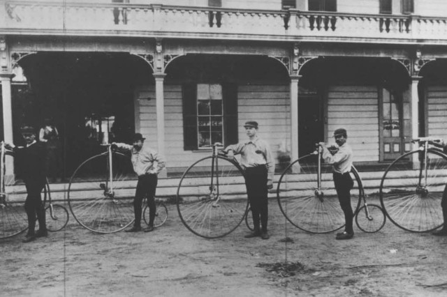 Antique photo of five men standing sideways in front of a building with a covered porch. They are each posing next to a high wheel antique bicycle.