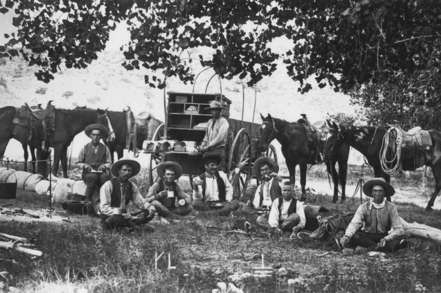 Antique photograph of eight cowboys sitting on the grass, eating a meal, under that shade of a large tree, with a chuck-wagon and four saddled horses behind them.