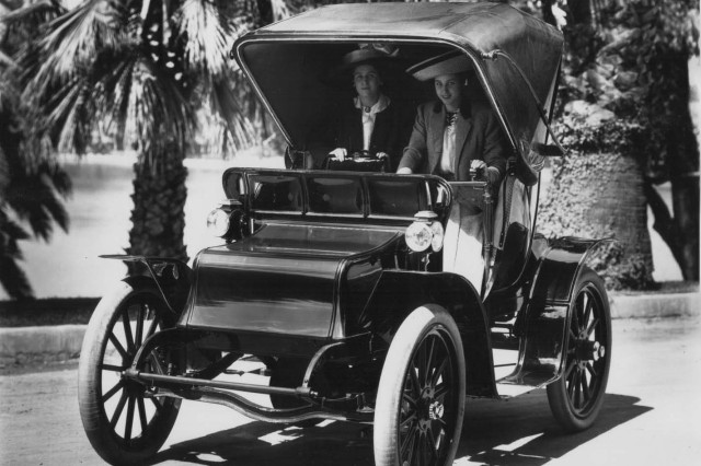 Antique photo of two ladies under the awning of a motorized carriage in front of a row of palm trees next to the road