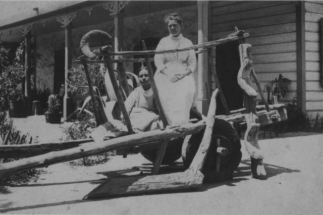 Antique photo of two pioneer women sitting in a wooden oxcart, one sitting on her heels and the other sitting on a chair, in front of an adobe house with a covered parch and partial siding