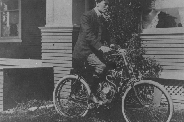 Antique photograph of a young man posing on a motorized bicycle parked on the grass in front of a house with a large porch. The man wears a driving hat, sports jacket over a white collared shirt, dark knee-high socks over stripped pants, and leather shoes.