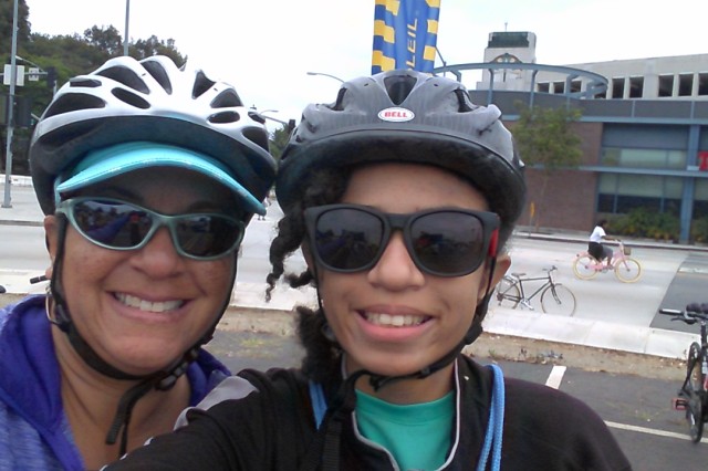 Selfie of a smiling mother and daughter of color, both wearing sunglasses and bike helmets.