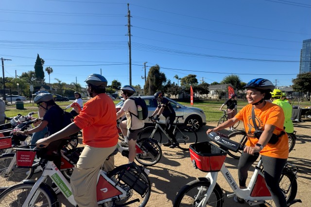 A dozen BIPOC bicycle riders, on both normal and electric bicycles, riding on a dirt path past a car parked next to a large grassy lawn with trees below a powerline in the background.