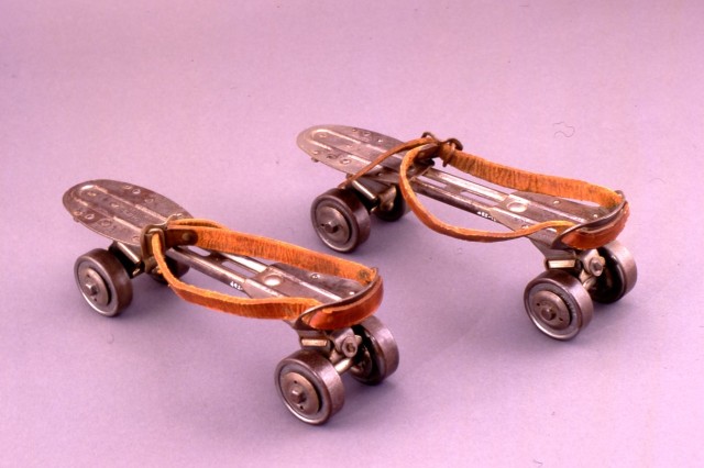 Antique strap-on roller skates on a pink background. Flat metal soles with four small metal wheels below and a leather strap on top to slip a show into.