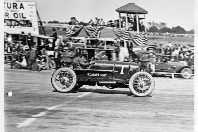 Antique photograph of early open-top, two-seater race car with driver and passenger passing by a blurred grandstand surrounded by a large crowd of people.