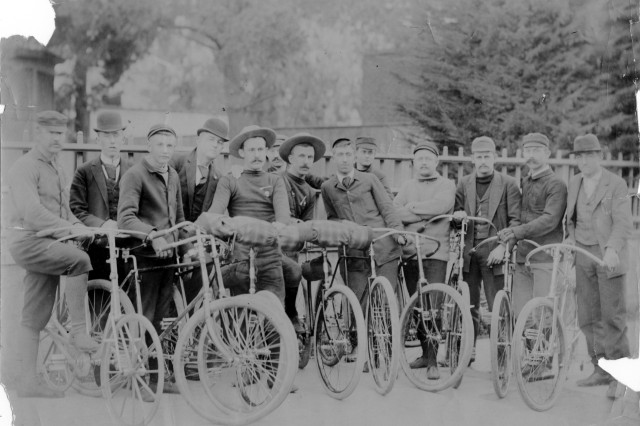 Antique photograph of 13 men lined up side by side astride their bicycles. The mean wear hats, sport coats, short pants and knee-high socks with leather shoes..