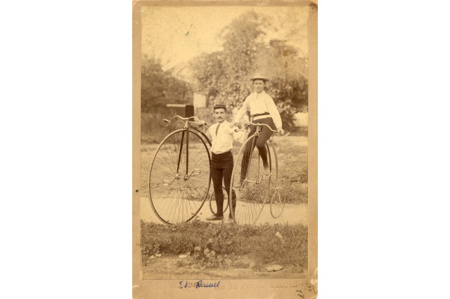 Antique photograph of two men with high wheel bicycles (large front wheel and small back wheel). Both men wear white shirts with ties, black tights and leather shoes. The mustached older man on the left stands in between the two bikes with his hands on the handlebars, the younger man sits astride the bike on the right.