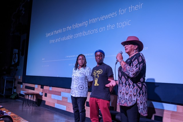 A BIPOC man and woman standing on stage in front of a movie screen while a moderator speaks into a microphone.