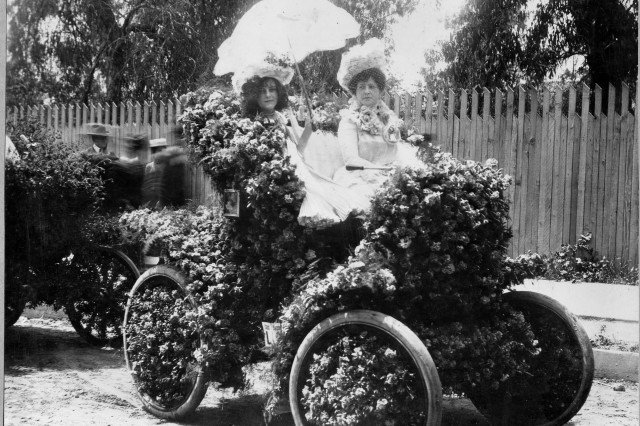 Antique photo of a motorized carriage covered in flowers, with two women dressed in white dresses, one holding a parasol.