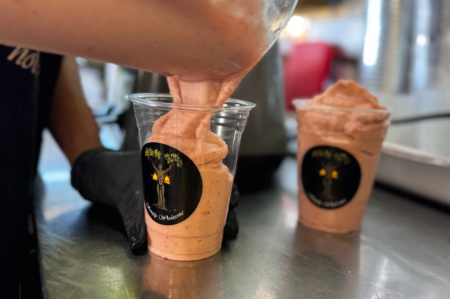 Mia Keeling smiling as she holds up the finished "Yes We Can" smoothie in a clear cup, with the Simply Wholesome logo visible.