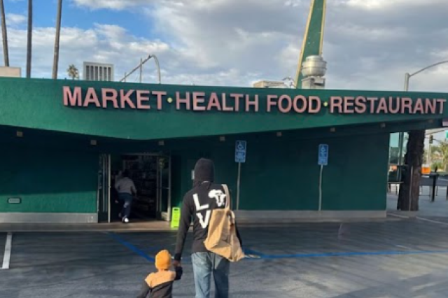 A view from behind of a small child in an orange beanie and an adult walking hand-in-hand toward the entrance of Simply Wholesome. The dark green building features a large sign reading "MARKET • HEALTH FOOD • RESTAURANT" across the top.