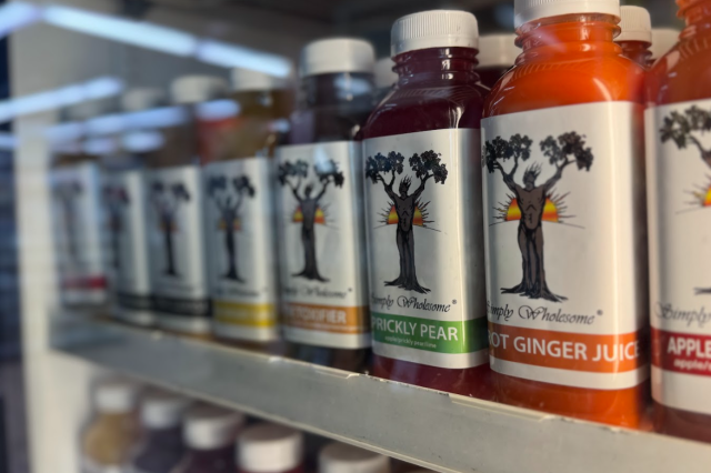 A row of colorful, bottled Simply Wholesome juices on a refrigerated shelf, including "Prickly Pear," "Carrot Ginger Juice," and "Apple Berry."