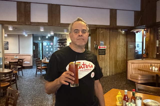 A man stands in a diner wearing a Canter's shirt hold a glass with a bloody mary 