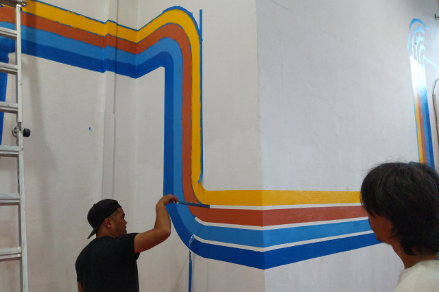 A man peels paint off a wall between 4 colored lines winding on a white wall in the storefront.