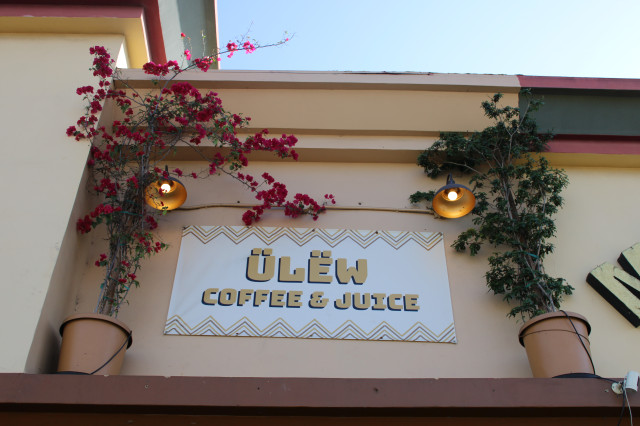 Ulew Coffee & Juice is on a sign above a storefront. The sign is framed by greenery and paperflower.