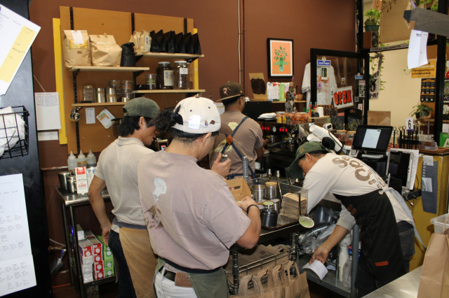 Four men work through the cafe making drink orders, packing up drinks, cleaning the workspace and taking orders. 