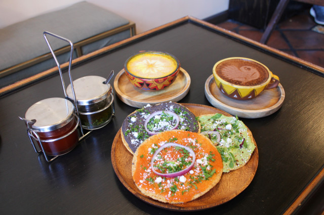 An orange plate of three tostadas sits next to two drinks from Ulew, and some condiments on a wooden table
