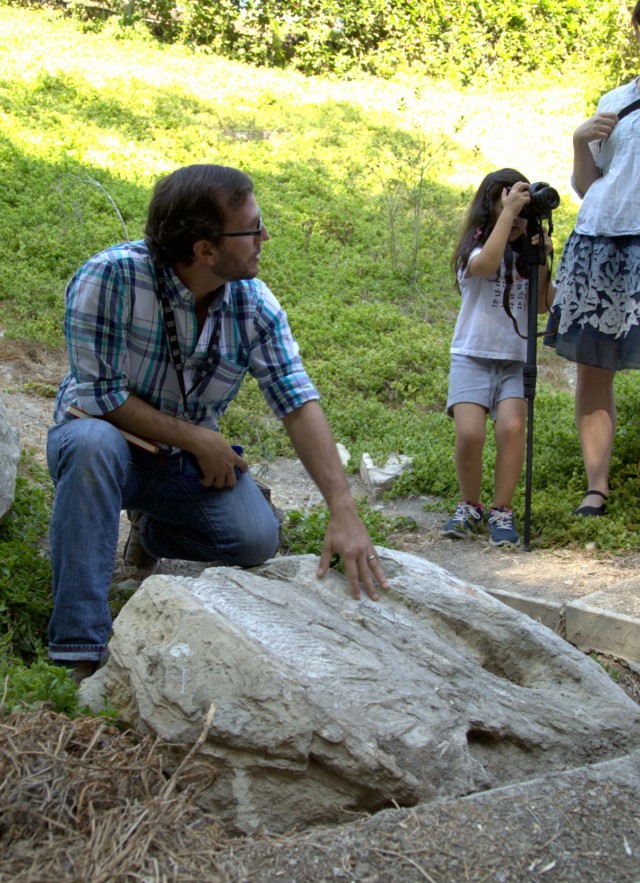 Man in plaid shirt standing over rock. Child and woman in background 