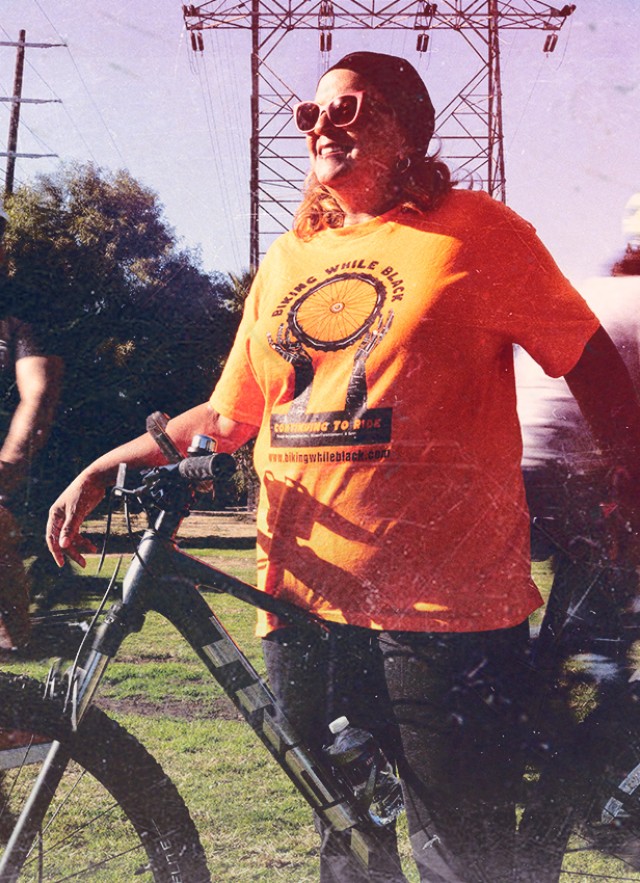 Woman of color in an shirt with Biking While Black logo on it, standing astride a bicycle with other cyclists behind her on a grassy lawn.