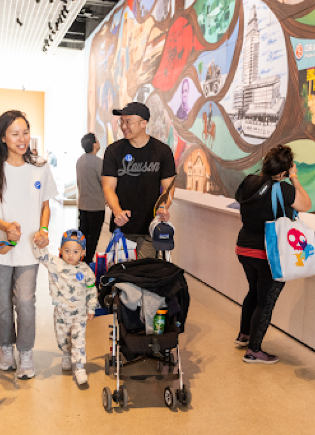 Family of man pushing stroller and woman holding toddler and older child&#039;s hands walking past Carasco mural in NHM Commons