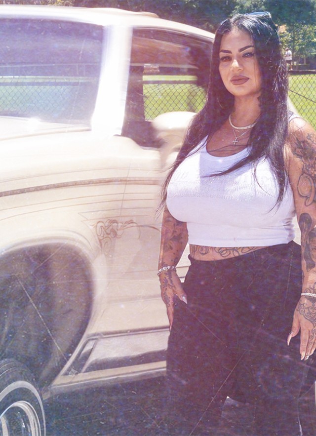 Latino woman in a white tank top and black capris standing next to a white lowrider car.