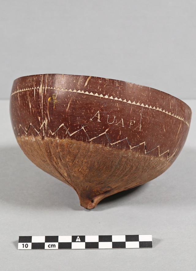 A wide-angle, eye-level studio photograph shows a deep, rounded bowl crafted from a polished coconut shell. The bowl is tilted slightly to the right, resting on a small, pointed nub at its base. It sits against a neutral, light-gray background. A black-and-white centimeter scale bar is positioned in the foreground for size reference.