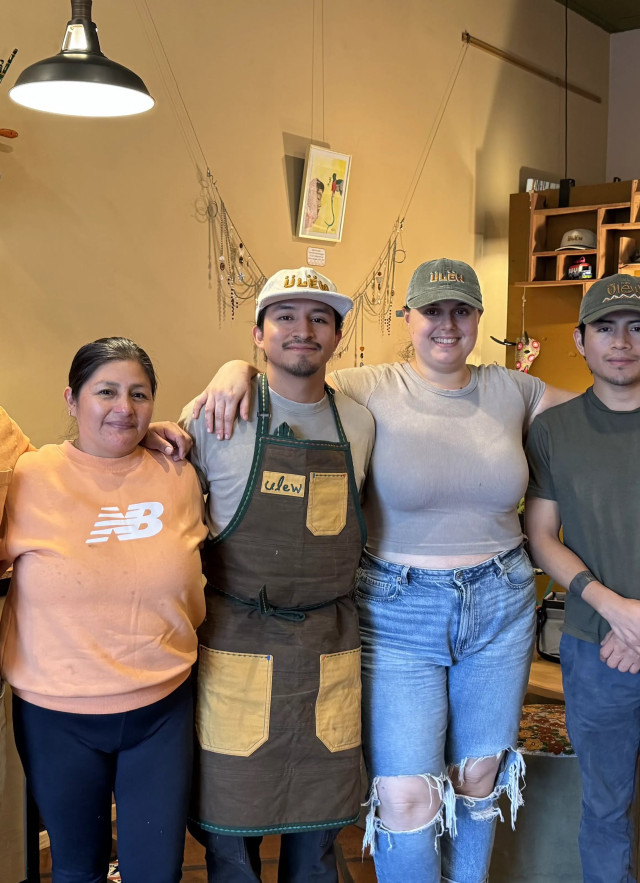 A family portrait of cafe owners Rosa and her son Jefri, standing with Jefri's partner Michaela, uncle Elvis, and brother Christopher. They are smiling in the cafe's cozy seating area, which features warm ambient lighting, vibrant house plants, and artwork on the walls.