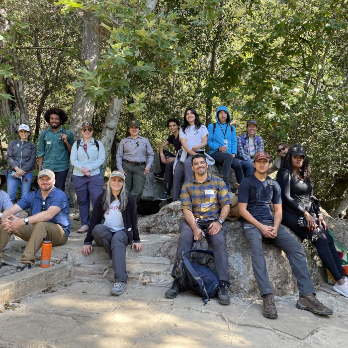 Group of students, NHM employees, and nature guide sitting and standing with trees in the background