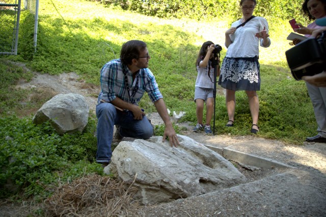 Man in plaid shirt standing over rock. Child and woman in background 