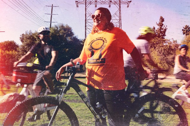 Woman of color in an shirt with Biking While Black logo on it, standing astride a bicycle with other cyclists behind her on a grassy lawn.