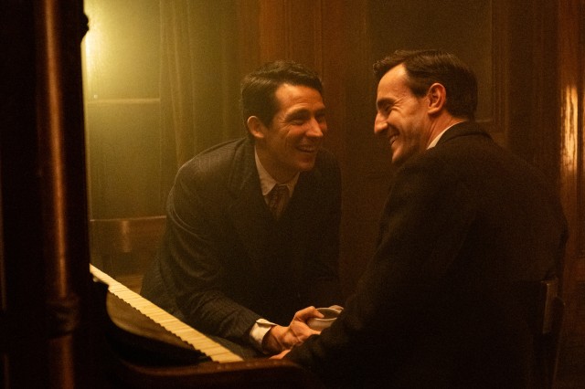 Two men in suits sitting by a piano, facing each other