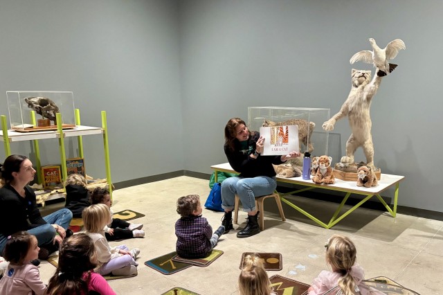 A Museum Educator is reading a book to a group of young children seated on the floor in an educational exhibit space. The room contains display cases with skulls and taxidermied animals, including a large wild cat is catching a bird. Stuffed animal toys are also visible near the reader.