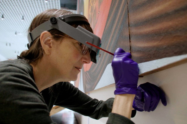 Tania Collas wearing magnifying head piece and purple gloves while holding a small paintbrush up to a mural