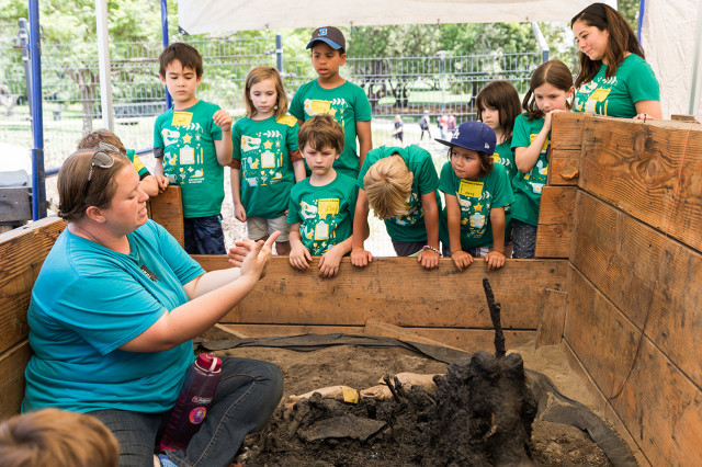 Excavator sitting by a fossil at an excavation site, surrounded by children looking on