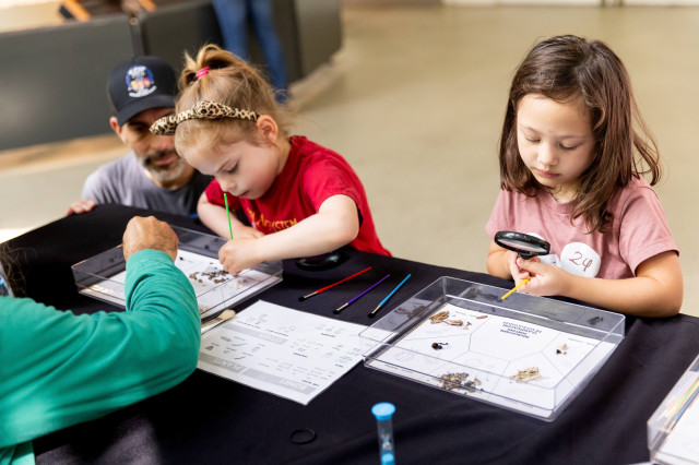 Two young girls at a black table working on microfossil sorting with paintbrushes and a magnifying glass