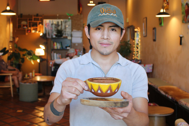 Man in baseball cap holding out a colorful tea cup inside a cafe.