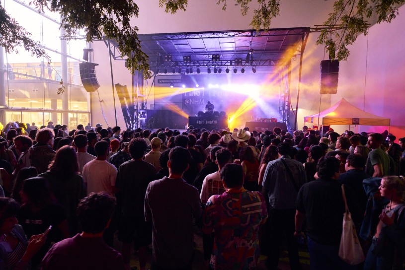 Outdoor nighttime scene of a majority seated audience in an amphitheater, surrounded by trees, facing a DJ stage that is emanating colorful fluorescent lights, with the Natural History Museum building in the background. 