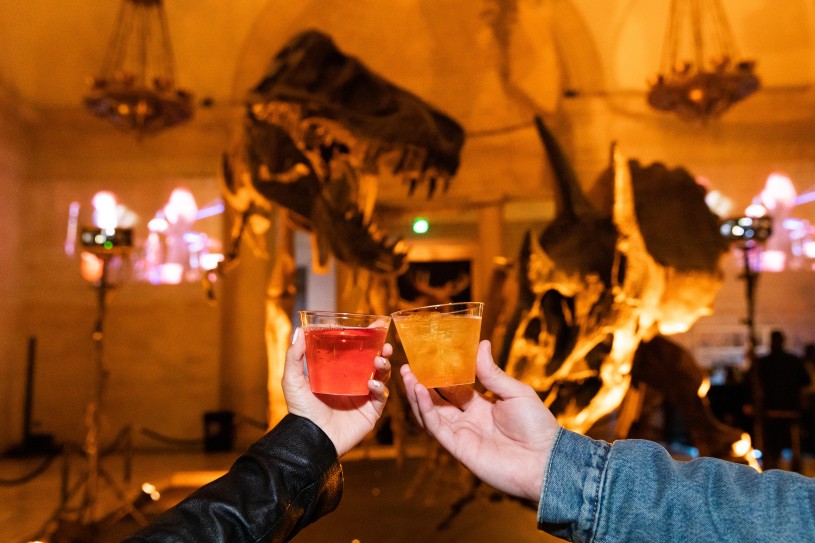 Two guests bring their glasses together for a cheers with Dueling Dinos in the background. One hand is holding a red drink and the other hand is holding an orange drink.