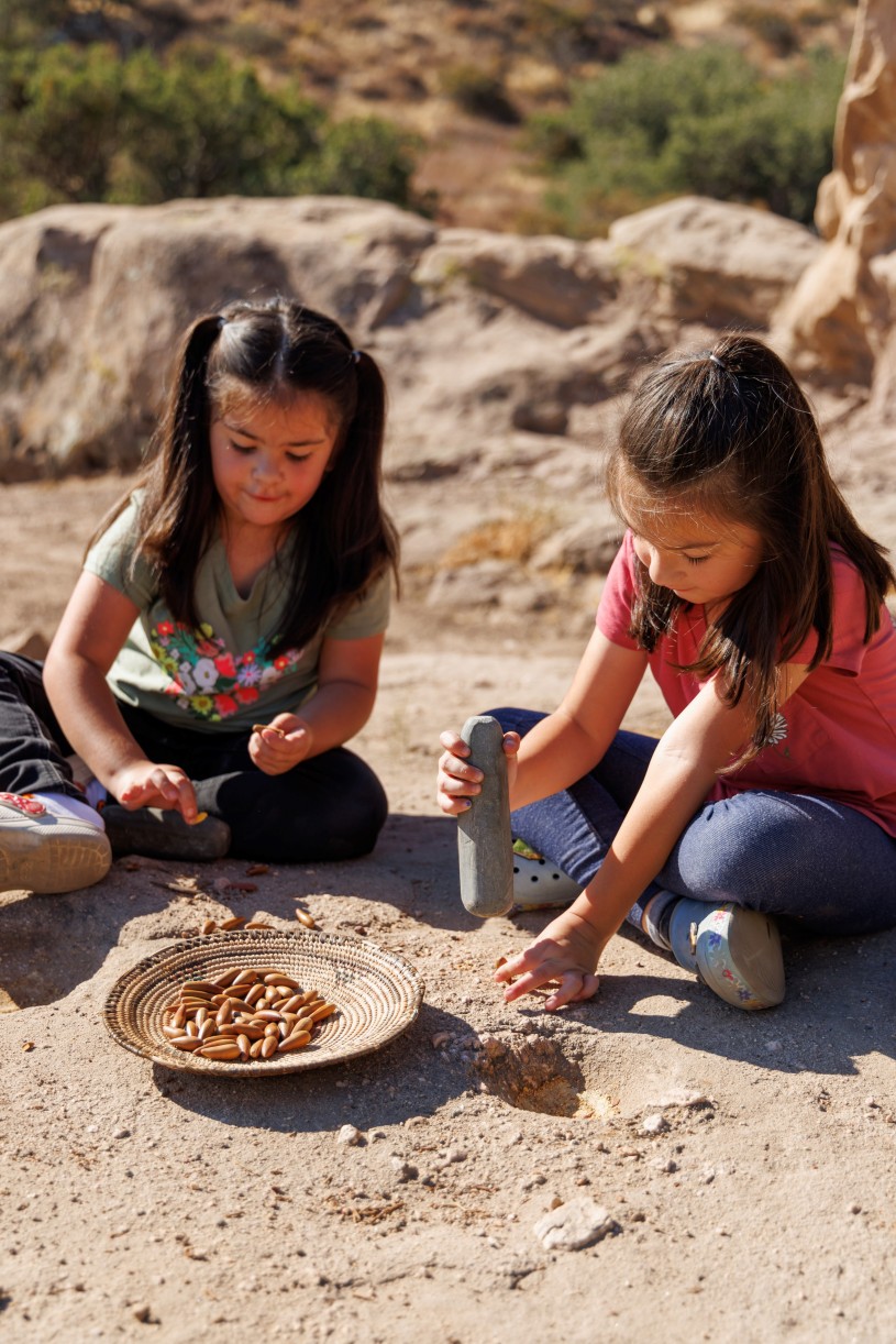 Two children from the Fernandeno Tataviam Band of Mission Indians sit atop a bedrock mortar. They use pestles to grind acorns and a woven basket with acorns sits at their feet. 