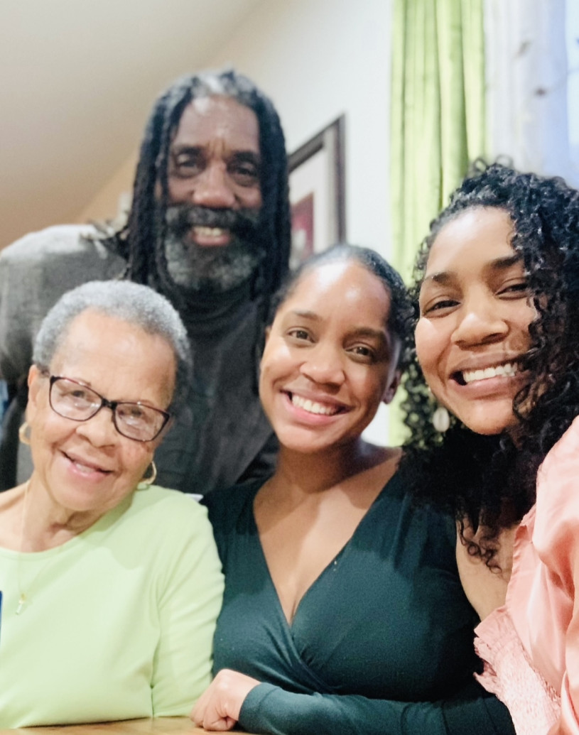 Four people of African descent smile warmly in a close-knit group portrait that radiates family joy. In the foreground, three women are gathered together: an older woman in a lime-green shirt and glasses on the left, a younger woman in a dark teal V-neck in the center, and a woman with voluminous dark curls in a peach ruffled top on the right. Standing protectively behind them is a man with long dreadlocks and a grey-flecked beard, looking directly at the camera with a kind expression. 