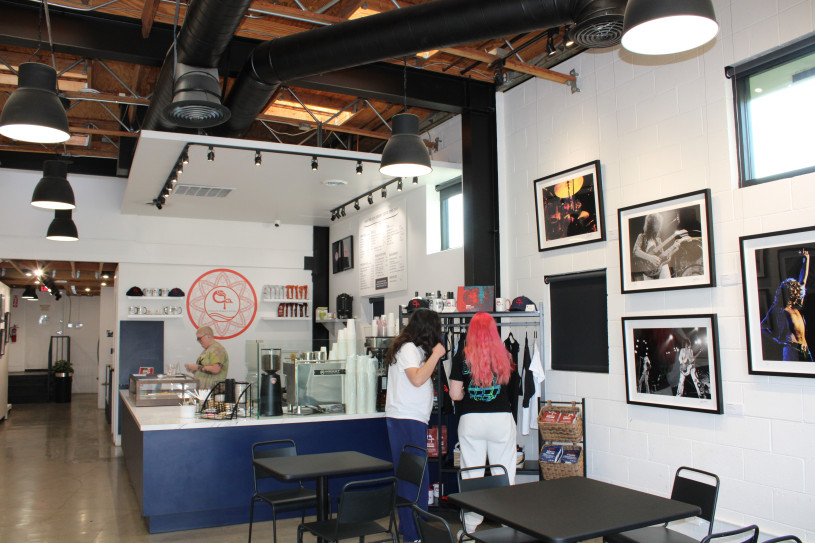 Pictured is the interior of the coffee shop featuring an exposed ceiling with black vents, lighting, and wooden rafters. A blue counter wraps around the central workspace, in front of which merchandise is displayed on a rack. The walls are decorated with various photos of Jimmy Page performing. 