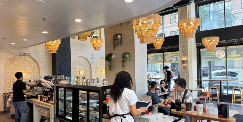 A wide shot of the interior of Sana'a Cafe, showing a bustling, vibrant atmosphere with diverse groups of people sitting at tables, engaged in conversation, and enjoying drinks together.
