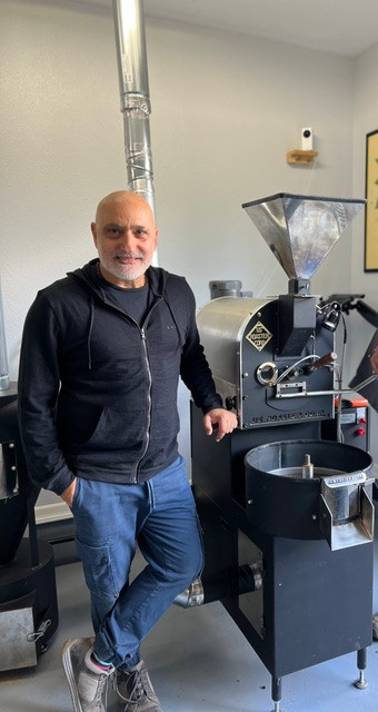 An adult Armenian man in a black zippered sweater and blue jeans stands next to a coffee bean roasting machine.